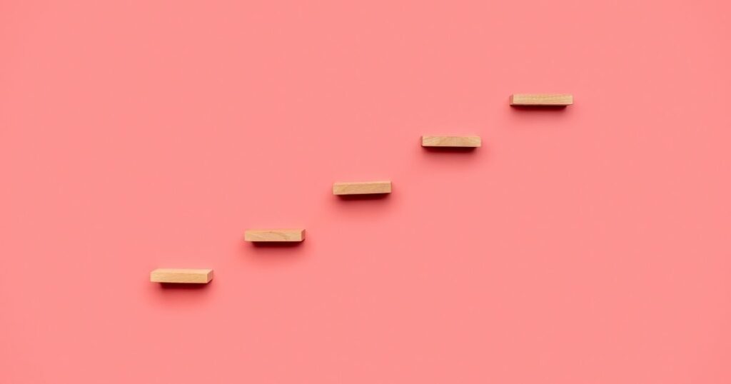 ピンク背景に置かれた階段状のブロック。願いを叶えるためのステップアップを象徴する構成。