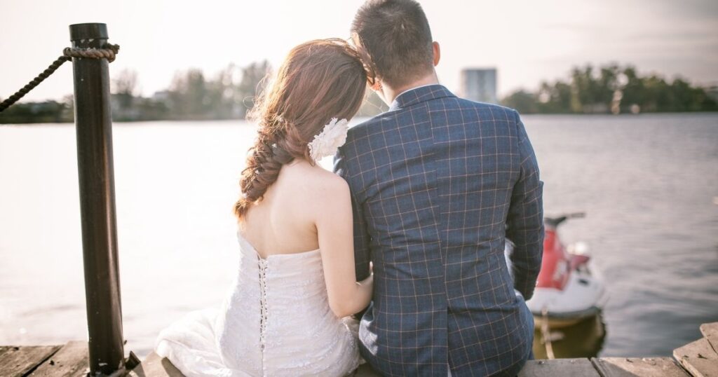 湖の桟橋で寄り添って座る夫婦の後ろ姿。結婚式風の衣装を着た女性とスーツ姿の男性。