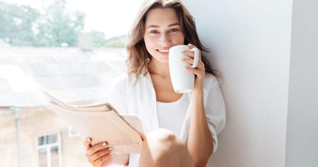 窓辺で笑顔でコーヒーカップを持つ女性。自然光が差し込み、リラックスした表情をしている。